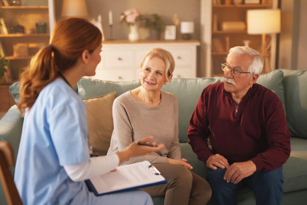 A nurse in scrubs, holding a clipboard, talks to an elderly couple on a sofa in a warmly lit living room. The couple listens attentively, conveying trust.