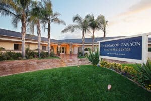 Front view of Cottonwood Canyon Healthcare Center, featuring a well-maintained garden with palm trees and a prominent center sign against a sunset sky.