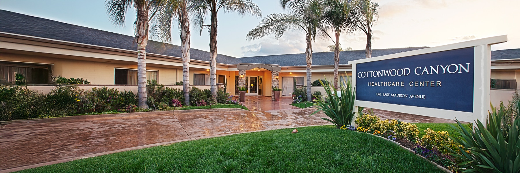 Front view of Cottonwood Canyon Healthcare Center, featuring a well-maintained garden with palm trees and a prominent center sign against a sunset sky.