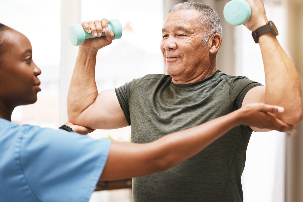 PACS-300x200-rehab2 An older man using light blue dumbbells is assisted by a healthcare worker in a blue uniform.