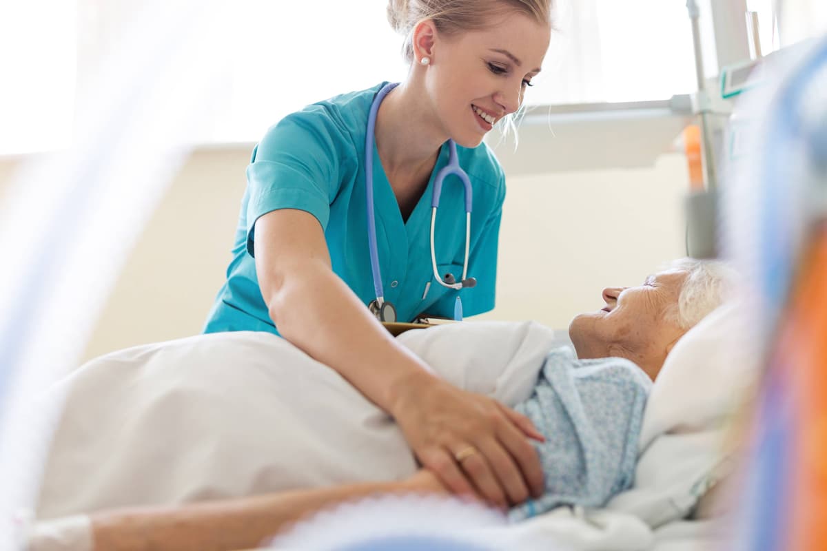 PACS-300x200-caregiver4 A nurse in blue scrubs, smiling warmly, cares for an elderly patient in a hospital bed.