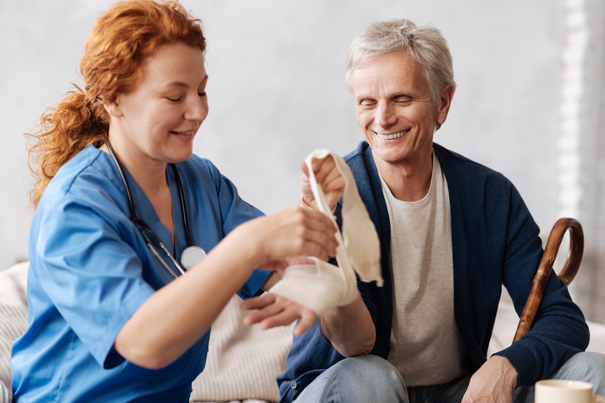 PACS-300x200-woundcare2 A smiling nurse with red hair in blue scrubs gently bandages the arm of an elderly man holding a cane, conveying a sense of care and trust.