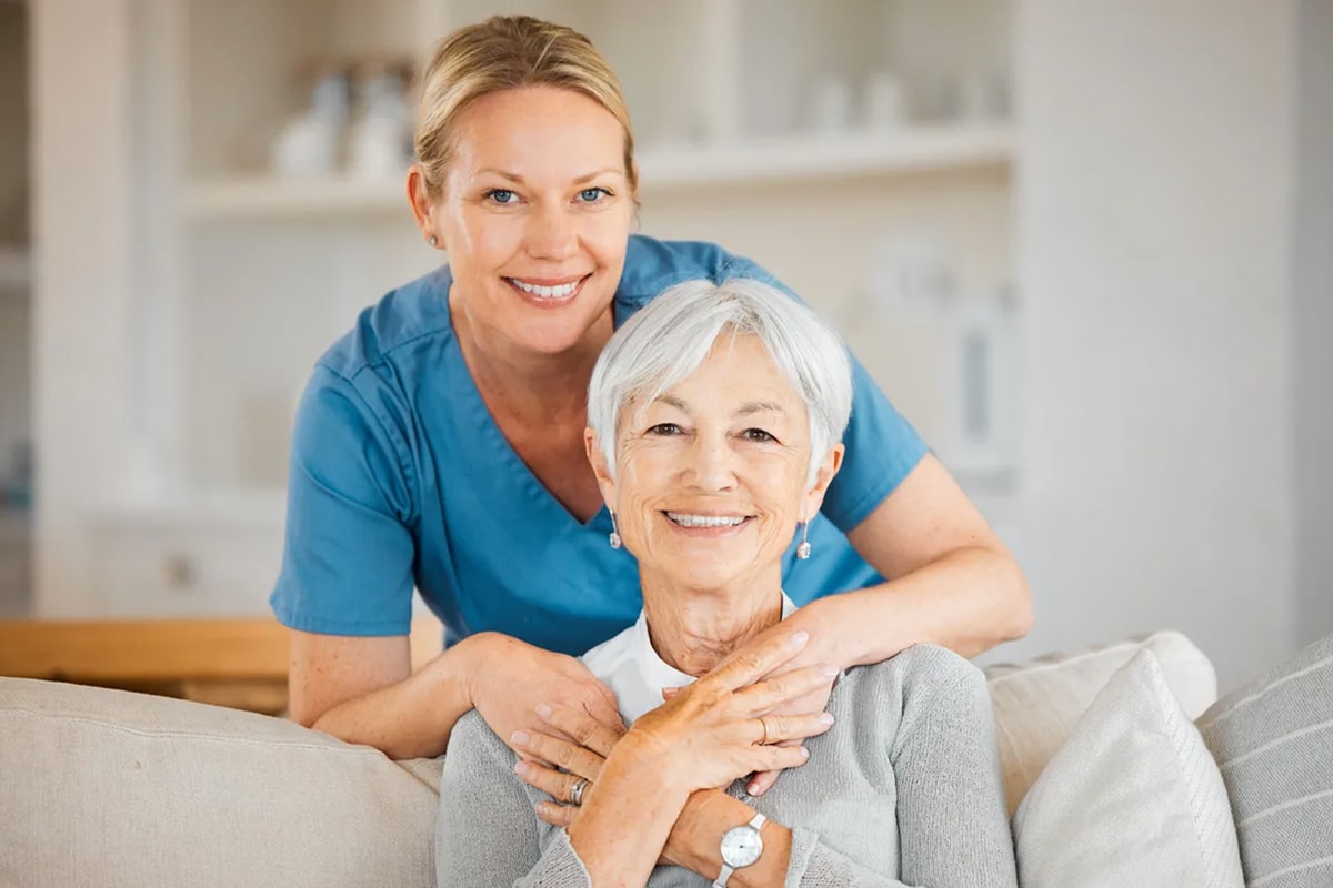 PACS-300x200-caregiver3 A smiling elderly woman with gray hair sits on a sofa, embraced from behind by a woman in a blue uniform.