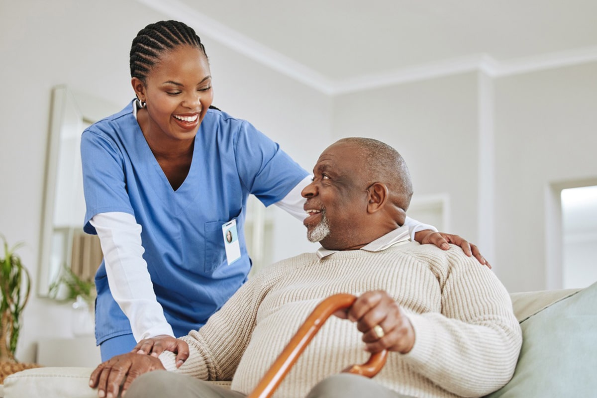 PACS-300x200-caregiver2 A smiling nurse in blue scrubs supports an elderly man holding a cane. They share a warm, friendly interaction in a cozy living room setting.