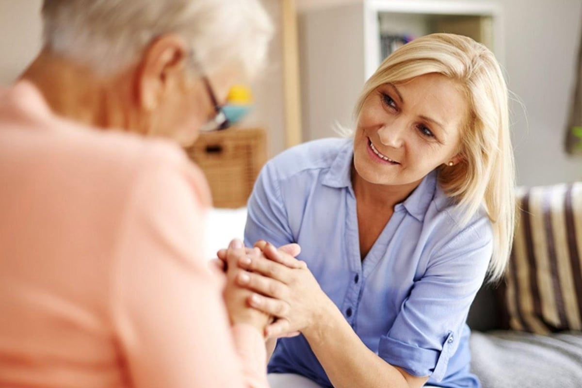 PACS-300x200-caregiver1 A woman in a blue shirt holds hands with an elderly person in peach, conveying empathy and warmth in a cozy, well-lit living room setting.