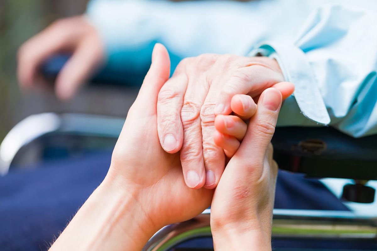 PACS-300x200-hands1 Close-up of young hands holding an elderly person's hand resting on a wheelchair, conveying care, support, and compassion in a bright setting.