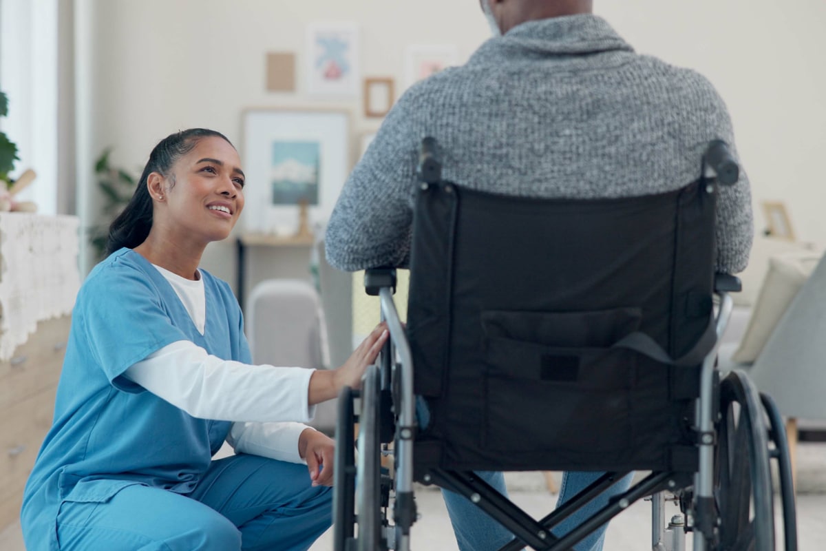 PACS-300x200-caregiver5 A nurse in blue scrubs kneels beside a person in a wheelchair, smiling and offering comforting support.