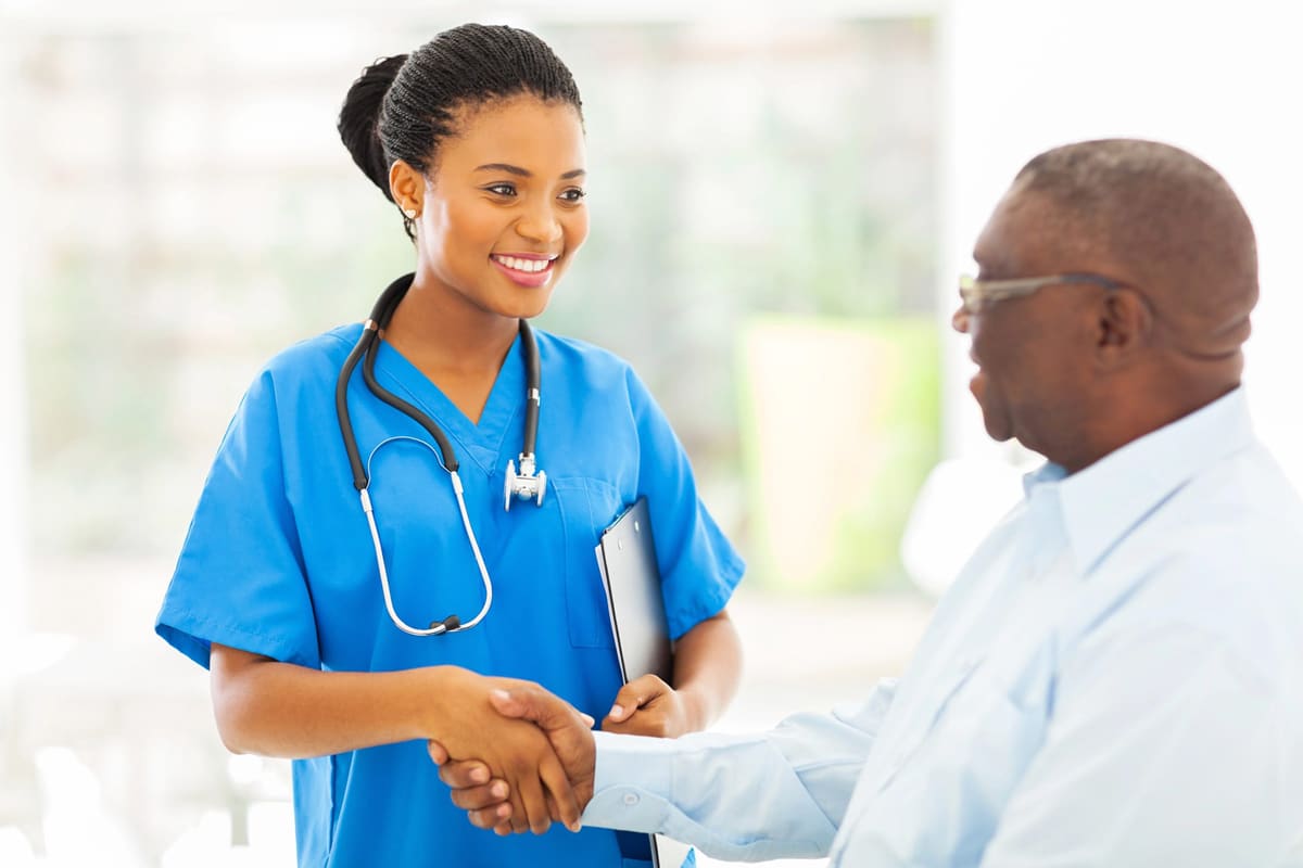 yolo-300x200-51 A smiling nurse in blue scrubs and stethoscope shakes hands with an older man in a light shirt.