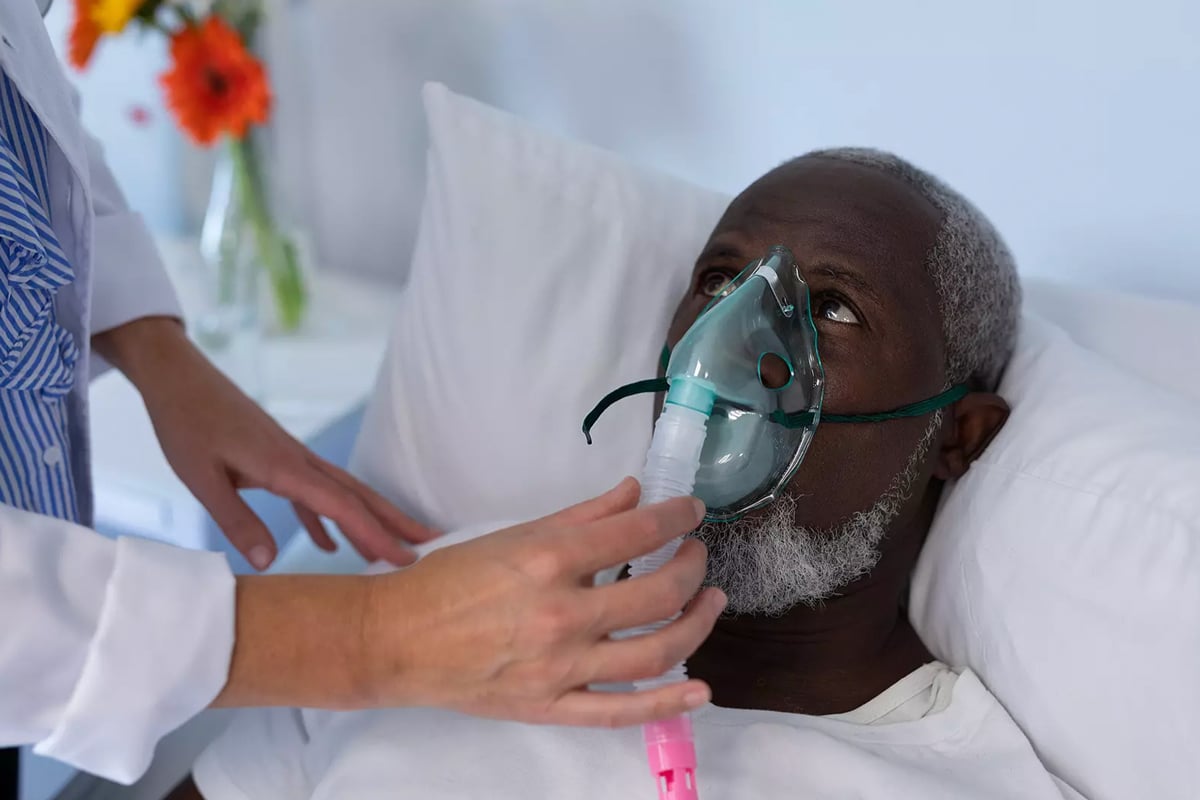 yolo-300x200-83 A man in bed wears an oxygen mask, assisted by a caregiver's hands. The setting is a hospital room with a calm atmosphere, flowers seen in the background.