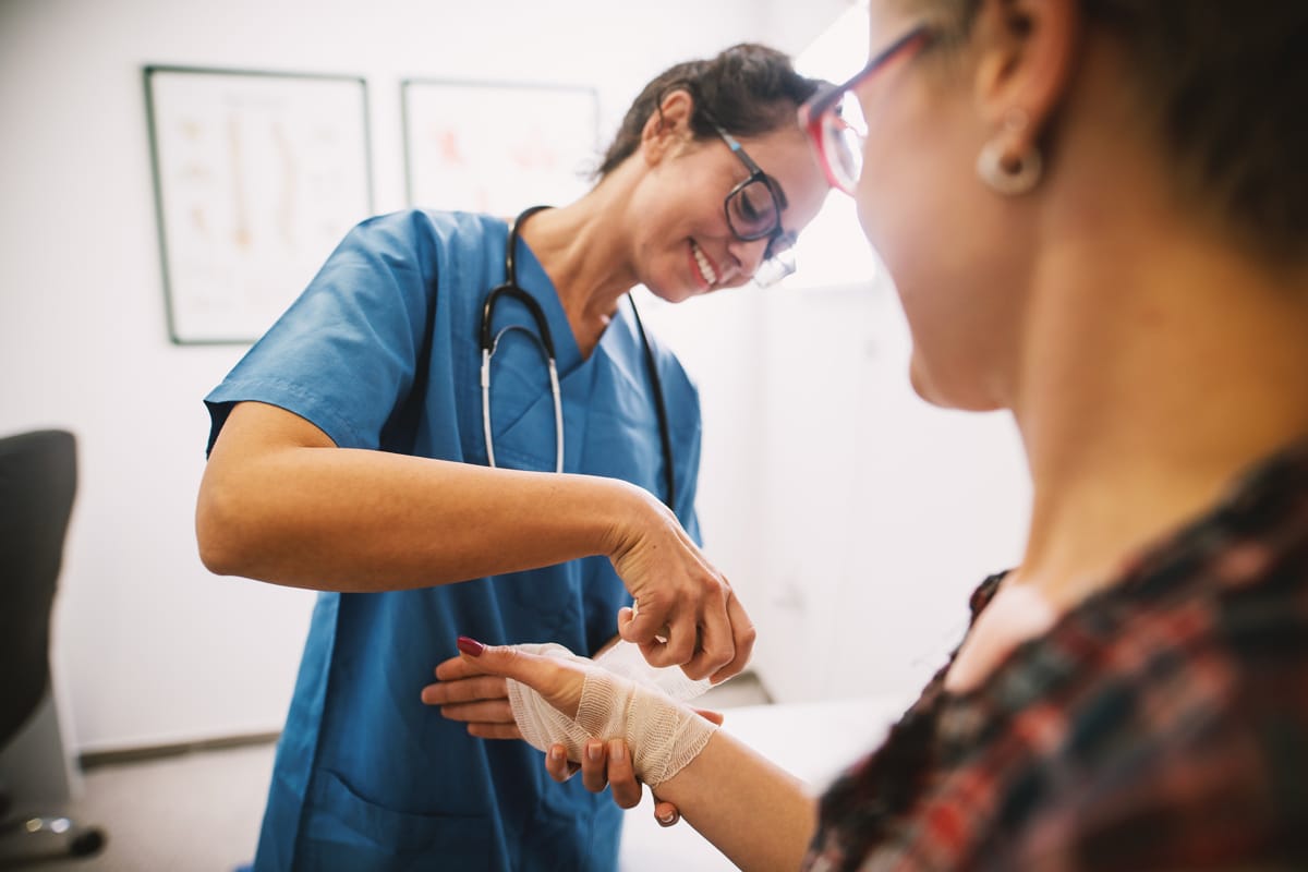 PACS-300x200-woundcare A smiling nurse wearing blue scrubs bandages a patient's hand in a medical office. The patient, wearing glasses, watches attentively.