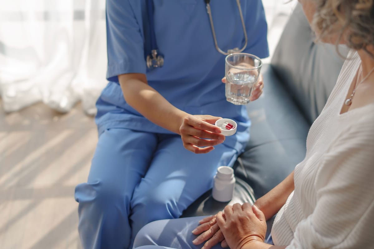 PACS-300x200-pain1 A nurse in blue scrubs offers pills and a glass of water to a seated patient in a cozy setting, conveying care and support.