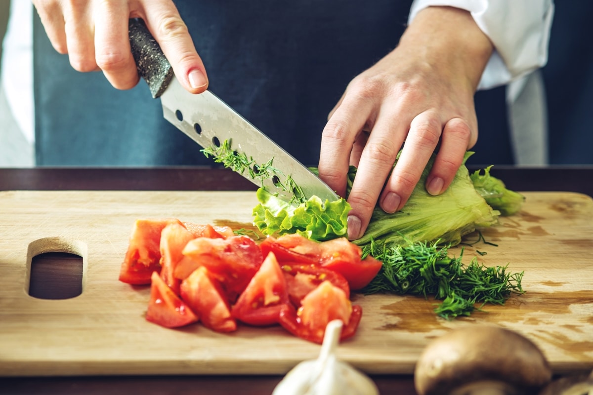 PACS-300x200-food1 Hands chopping lettuce and herbs on a wooden board with a knife, surrounded by diced tomatoes, mushrooms, and garlic.