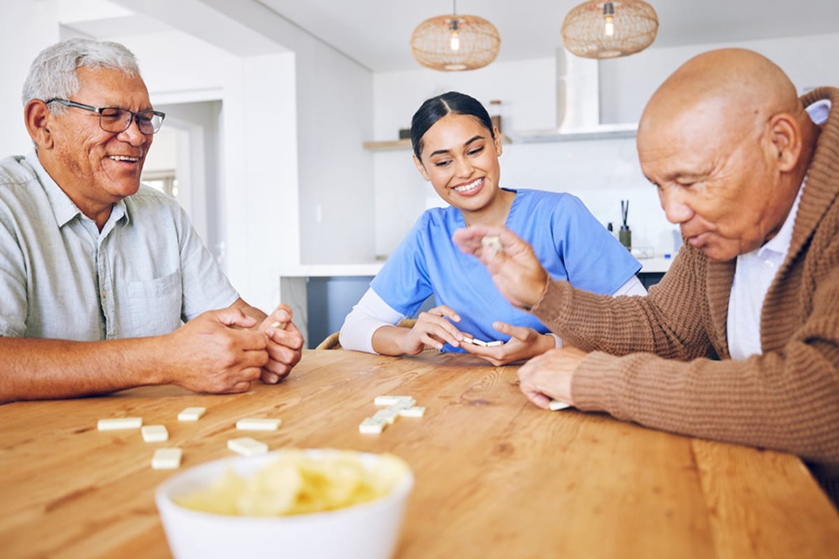 PACS-300x200-activity2 Three people enjoy playing dominoes at a wooden table. A smiling woman in blue scrubs sits between two elderly men. Warm lighting creates a friendly atmosphere.