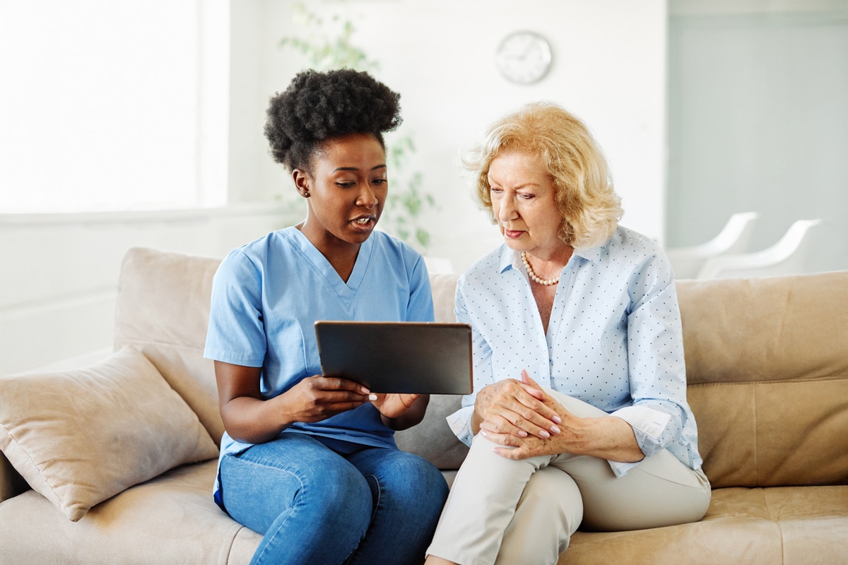 PACS-300x200-computer2 A nurse in blue scrubs sits with an elderly woman on a couch, discussing information on a tablet.