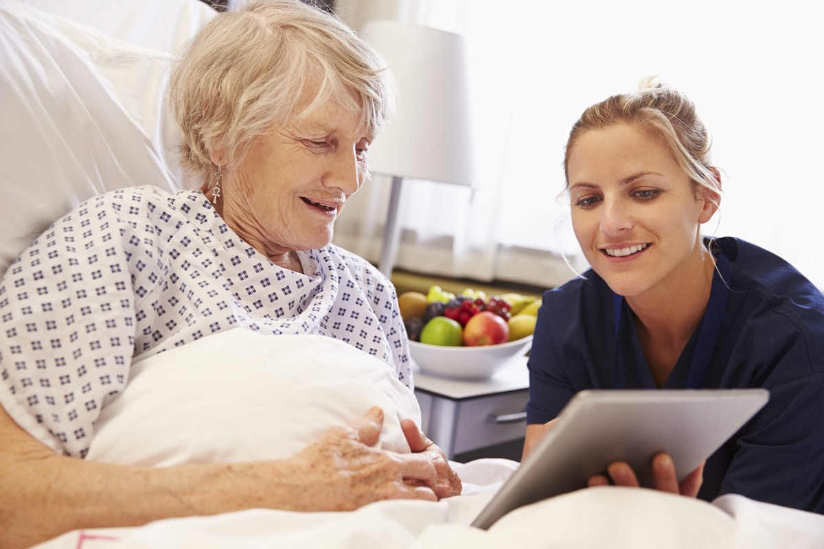 PACS-300x200-computer1 A nurse in blue scrubs shows an elderly woman in a hospital bed a tablet. They both smile warmly, creating a comforting and positive atmosphere.